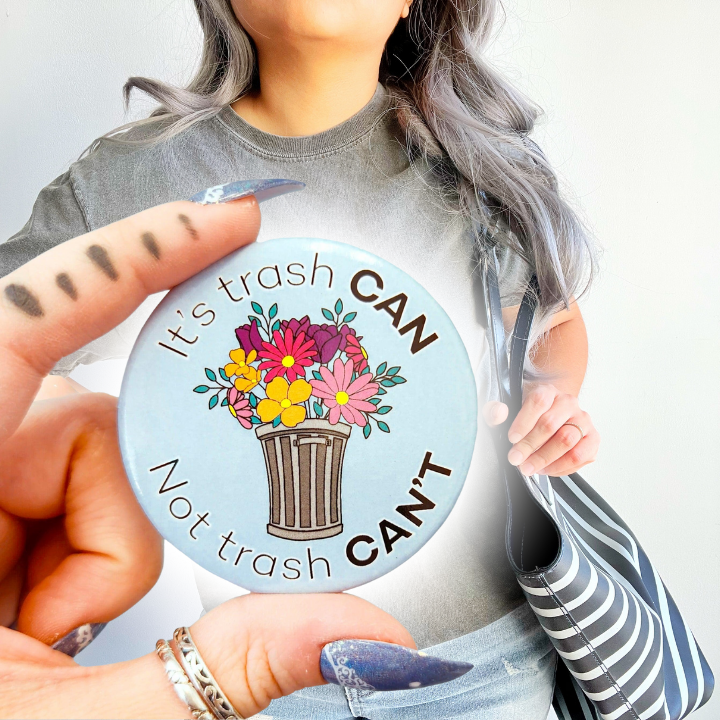 Person in a gray t-shirt holding a striped bag, with a closeup of a hand holding the "It's trash CAN Not trash CAN'T" motivational pin in the foreground.