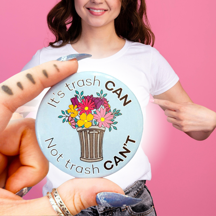 A smiling person in a white t-shirt stands against a pink background, pointing at a large motivational button pin that reads "It's trash CAN Not trash CAN'T," featuring a trash can with flowers.
