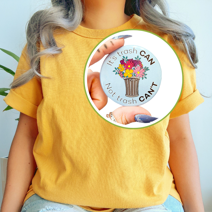 Person wearing a yellow t-shirt and light blue jeans, with a circular inset displaying the "It's trash CAN Not trash CAN'T" button pin being held up close.