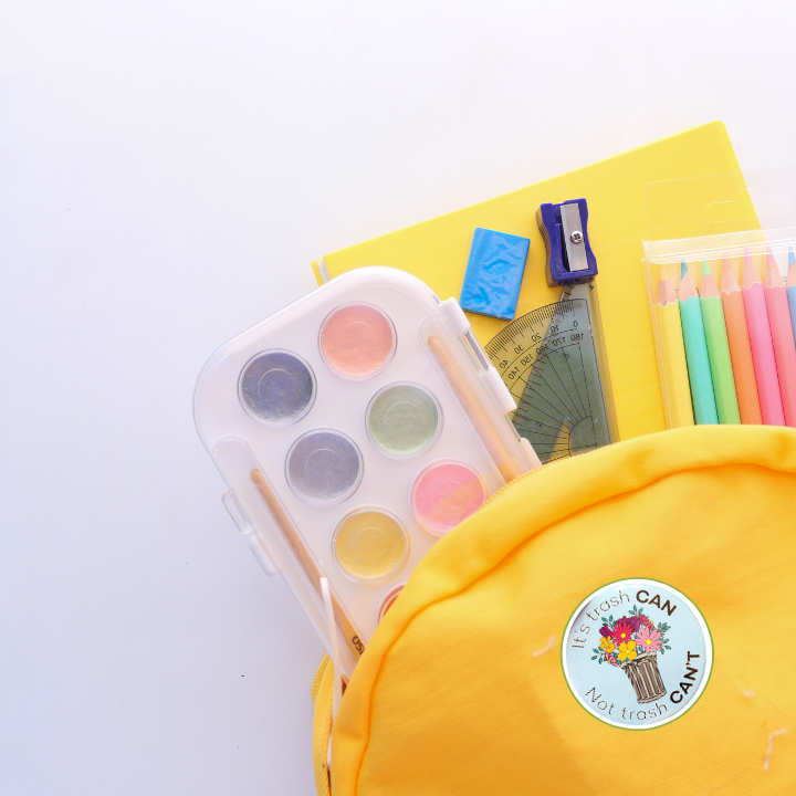 Flat lay of a yellow backpack surrounded by pastel school supplies, with the "It's trash CAN Not trash CAN'T" button pin attached to the backpack.